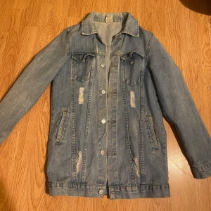 Blå jeansjacka - Snygg blå jeansjacka med klassisk design och slitna detaljer. Jackan har långa ärmar och knappar framtill samt två bröstfickor med lock. Perfekt för vår,höst och sommar.