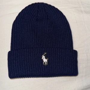 Mörkblå  Ralph Lauren mössa - Snygg mörkblå ribbad mössa från Polo Ralph Lauren med det ikoniska broderade logotypen framtill. Perfekt för att hålla dig varm och stilren under kyliga dagar.