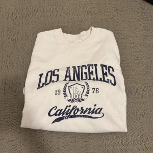 Vit sweatshirt från Pier One - Säljer en vit sweatshirt från Pier One med tryck 'Los Angeles California 1976' i blått. Tröjan har långa ärmar och en rund halsringning.