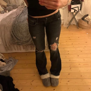 Svarta slitna jeans lågmidjade med hål - Snygga svarta jeans med slitningar på knäna och låren, bootcut och låg midja. Stl 34. Har sytt de utsvängda och lågmidjade
