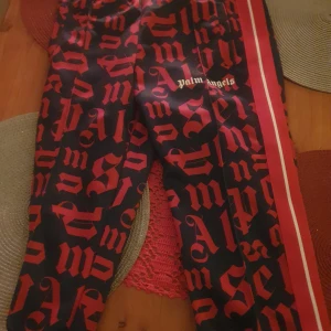 Svarta byxor med rött mönster från Palm Angels - Snygga svarta byxor från Palm Angels med ett iögonfallande rött gotiskt mönster. Byxorna har en röd och vit rand längs sidorna som ger en sportig touch. Perfekta för dig som vill sticka ut!