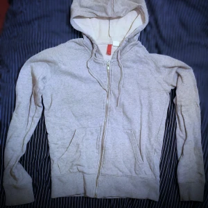 Grå hoodie med dragkedja - Säljer en bekväm grå hoodie med dragkedja och huva. Perfekt för en avslappnad stil. Den har långa ärmar och praktiska fickor framtill. Passar bra till både jeans och mjukisbyxor.