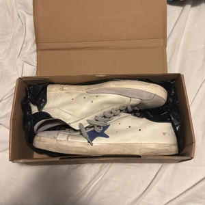 Vita sneakers med blå stjärna - Säljer ett par vita sneakers från Golden Goose med en blå stjärna på sidan. Skorna har grå snörning och en klassisk design. Perfekta för en avslappnad stil. Köpta i Italien för 465€ skulle säga skorna passar 44, 45 