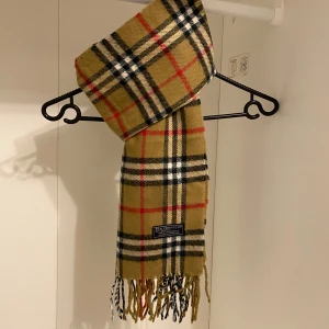 Burberry - Säljer denna snygg och klassisk rutig halsduk från Burberry i beige. Tillverkad i 100% mjukt lammull  med fransar i ändarna. Perfekt för att hålla dig varm och stilren under kyliga dagar. Hör av er vid minsta fundering☺️