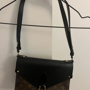 Monogram axelväska från Louis Vuitton - Snygg axelväska från Louis Vuitton med klassiskt monogrammönster. Väskan har en svart läderklaff och justerbar axelrem. Perfekt för att bära dina dagliga nödvändigheter med stil. Finns i Trollhättan 