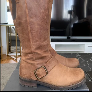 Bruna skinnstövlar med spänne - Snygga bruna stövlar i skinn med robust sula och dekorativt spänne. Perfekta för höst och vinter, dessa stövlar ger både stil och komfort.