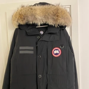 Svart parkajacka med päls från Canada Goose - Säljer en svart parkajacka från Canada Goose med pälsfodrad huva. Jackan har knappar och flera praktiska fickor framtill. Perfekt för kalla vinterdagar. Knappt använd "pris kan diskuteras"