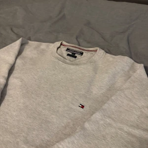 Grå tröja från Tommy Hilfiger - Säljer en stilren grå tröja från Tommy Hilfiger i mjuk bomull. Tröjan har en liten broderad logotyp på bröstet och rund halsringning med ribbade kanter. Perfekt för en avslappnad look.