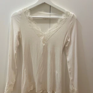 Vit långärmad topp med spetsdetaljer - Elegant vit långärmad topp från Intimissimi med v-ringning och spetsdetaljer vid hals och ärmslut. Perfekt för en stilren look. Säljer pågrund av att den aldrig kommit till användning. Toppen är i toppskick utan några defekter! Bara att skriva för närbilder på den och pris kan även diskuteras vid snabb äffär!💕