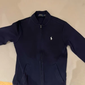 Mörkblå tröja  från Polo Ralph Lauren - Säljer en stilren mörkblå tröja  från Polo Ralph Lauren med dragkedja och broderad logga på bröstet. Tröjan  har långa ärmar och en klassisk design som passar perfekt för en avslappnad look.