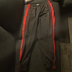 Svarta träningsbyxor med röda detaljer från Puma - Snygga svarta träningsbyxor från Puma med röda ränder längs sidorna. Byxorna har elastisk midja och är perfekta för sport och träning. Puma-loggan syns i blått på framsidan. Och de behöver inte vara tränings byxor de funkar också som hemma och om du är inte i sommaren 