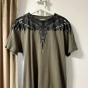 Olivgrön t-shirt med vingmönster från Marcelo Burlon - Snygg olivgrön t-shirt från Marcelo Burlon med ett coolt svart och vitt vingmönster runt halsen. Perfekt för en unik och stilren look.