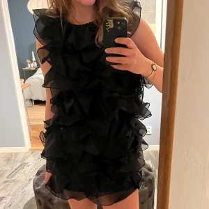 Svart volangklänning - Trendig svart klänning med flera lager av volanger som ger en lekfull och elegant look. Perfekt för en kväll ute eller en speciell tillställning. Klänningen är ärmlös och har en kort längd som framhäver benen.