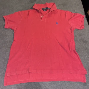 Röd pikétröja från Polo Ralph Lauren - Snygg röd pikétröja från Polo Ralph Lauren med klassisk krage och korta ärmar. Tröjan har en broderad blå logotyp på bröstet och är i en bekväm bomullskvalitet. Perfekt för en stilren look.