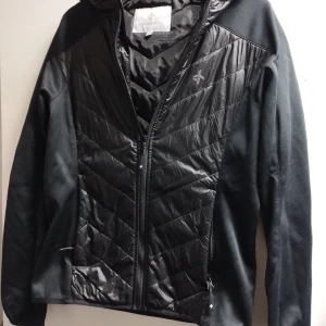 Svart jacka från Cross Sportswear - Snygg svart jacka från Cross Sportswear med quiltade detaljer och dragkedja. Perfekt för en sportig look med långärmad design och huva. Tillverkad i Sverige. Tunn sport eller vår/höstjacka 