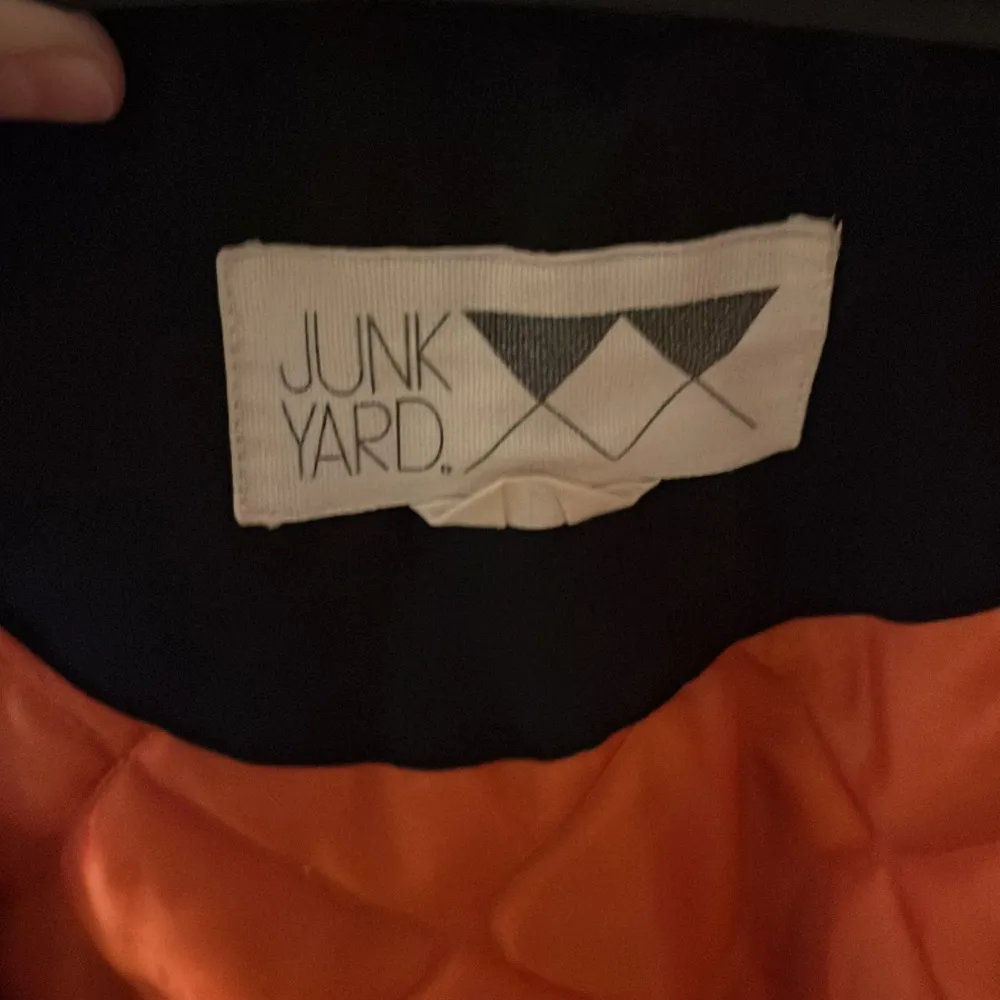 Snygg svart bomberjacka från Junkyard med orange quiltat foder. Jackan har dragkedja framtill och en ficka med dragkedja på ärmen. Perfekt för en cool och avslappnad stil.. Takit.