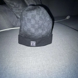 Svart rutig mössa från Louis Vuitton - Snygg svart mössa från Louis Vuitton med ett diskret rutigt mönster. Mössan har en uppvikt kant med en broderad logotyp framtill. Perfekt för att hålla stilen under kyliga dagar.
