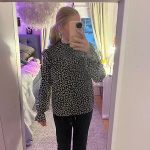 Svart blommig blus - Säljer en svart blus med vita blommönster. Blusen har långa ärmar och en hög hals. Perfekt för en stilren look.