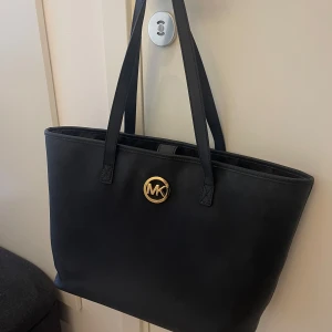Svart axelväska från Michael Kors - Snygg svart axelväska från Michael Kors med ett stilrent design. Väskan har ett guldigt MK-emblem på framsidan och långa handtag för bekväm bärning. Perfekt för att bära med sig det nödvändigaste med stil.