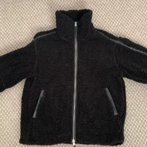 Svart teddyjacka från Hm - Mysig svart teddyjacka med dragkedja framtill och hög krage. Perfekt för kyliga dagar när du vill hålla dig varm och bekväm. Jackan har två fickor med svarta detaljer.
