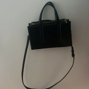 Svart axelväska från Zara - Snygg svart axelväska från Zara i skinn med flera dragkedjor och praktiska fack. Den har både handtag och en justerbar axelrem för flexibel användning. Perfekt för att hålla ordning på dina saker med stil.