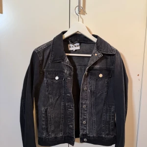Svart jeansjacka från NA-KD - Snygg svart jeansjacka från NA-KD med klassisk design. Jackan har långa ärmar och knappar framtill samt på bröstfickorna. Perfekt för en casual look.
