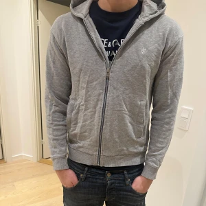 Grå marc o’polo hoodie - Säljer en stilren grå hoodie med dragkedja och huva. Tröjan har långa ärmar och en liten logga på bröstet. Perfekt för en avslappnad look.