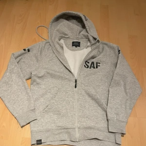 Grå hoodie med tryck från Swedish Armed Forces - Säljer en grå hoodie med dragkedja och tryck från Swedish Armed Forces. Har även en grön och en rosa