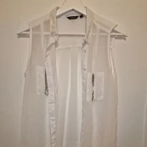 Vit ärmlös blus från Lindex - Snygg vit ärmlös blus från Lindex i ett transparent material. Den är dekorerad med kedjedetaljer på fickorna, liten defekt på den ena fickan därav lågt pris.