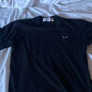 Svart t-shirt från Comme des Garçons - Säljer en svart t-shirt från Comme des Garçons med det ikoniska hjärtmärket med ögon på bröstet. T-shirten har en klassisk passform och är kortärmad. Perfekt för en stilren look.