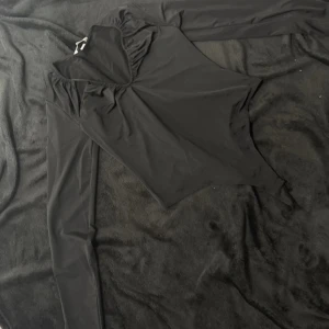 Svart långärmad body - Säljer en elegant svart långärmad body med hög krage och rynkdetaljer vid axlarna. Perfekt för en stilren look. Passar bra till både jeans och kjol.