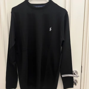 Svart långärmad tröja från Polo Ralph Lauren - Snygg svart långärmad tröja från Polo Ralph Lauren med det klassiska logotypbroderiet på bröstet. Perfekt för en stilren look. Tröjan har en rund halsringning och är tillverkad i ett mjukt material.