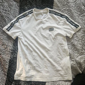 Vit t-shirt från Emporio Armani - Säljer en stilren vit t-shirt från Emporio Armani med EA7-logga på bröstet och längs ärmarna. T-shirten är kortärmad och tillverkad i mjukt bomullsmaterial. Perfekt för en avslappnad look.