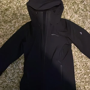 Svart jacka från Peak Performance - Snygg svart jacka från Peak Performance med huva och dragkedja. Perfekt för kyligare dagar med sin stilrena design och funktionella detaljer. Jackan har en liten logotyp på ärmen och är tillverkad i ett slitstarkt material.