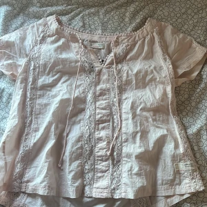 Ljusrosa blus från Odd Molly - Charmig ljusrosa blus från Odd Molly med vackra spetsdetaljer och knytband framtill. Blusen har korta ärmar och en avslappnad passform, perfekt för en bohemisk look. Den broderade loggan på baksidan ger en extra touch av stil. Aldrig använd då den inte är min stil. PRIS KAN DISKUTERAS!💕
