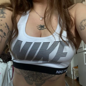 Vit sporttopp från Nike - Snygg vit sporttopp från Nike med svart logga och elastiskt band med texten 'NIKE PRO'. Perfekt för träning med sin bekväma passform och stödjande design.