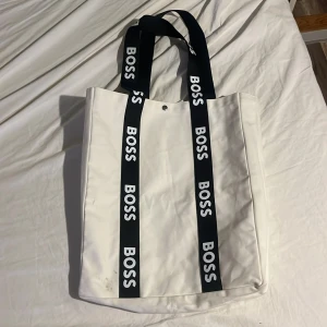 Vit axelväska från Boss - Snygg vit axelväska från Boss med svarta handtag och logotyp. Perfekt för att bära med sig det nödvändigaste med stil. Väskan har en enkel och modern design.