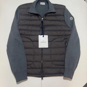 Grå jacka från Moncler - Snygg grå jacka från Moncler cardigan med quiltad framsida och stickade ärmar. Jackan har en dragkedja framtill och Moncler-logga på ärmen. Perfekt för kyligare dagar. Jackan passar M också eftersom den är tajt hos midjan. Priset kan diskuteras och jackan är perfekt kvalité och aldrig använd. 