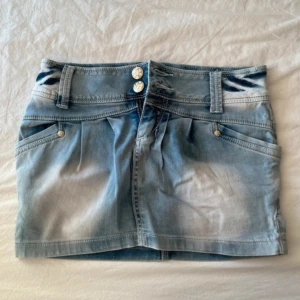 Blå jeanskjol med knappar - Snygg blå jeanskjol med två dekorativa knappar framtill och fickor både fram och bak. Passar perfekt nu till sommaren!! 