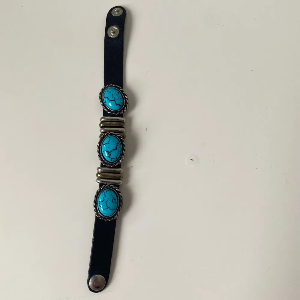 Snyggt svart armband med tre stora turkosa stenar infattade i silverfärgade detaljer. Ej äkta turkos. Armbandet har en justerbar tryckknappslåsning och metalliska dekorationer i guld och silver. Perfekt för att ge en bohemisk touch till din stil.. Asusteet.