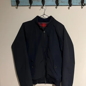 Mörkblå jacka från Baracuta - Säljer en stilren mörkblå jacka från Baracuta med klassiskt röd- och grönrutig insida. Jackan har dragkedja och knappar framtill samt långa ärmar. Perfekt för en snygg och avslappnad look. Hör gärna av dig! Pris kan diskuteras.