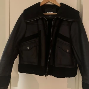 Svart jacka från DKNY Jeans - Snygg svart jacka från DKNY Jeans med dragkedja och två fickor med knappar framtill. Jackan har en stilren design med en krage i mjukt material och ribbade muddar. Perfekt för en cool och avslappnad look.