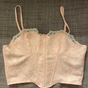 Rosa korsett topp med spetsdetaljer - Söt rosa korsett med blommigt mönster och spetsdetaljer längs kanten. Den har justerbara axelband och en dragkedja bak. 