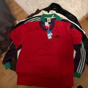 Röd t-shirt från Adidas - Säljer en snygg röd t-shirt från Adidas med det klassiska trefoil-logotypen på bröstet. T-shirten har en kontrasterande krage i lila och grönt. Perfekt för en sportig look!