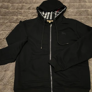 Svart hoodie från Burberry - Säljer en stilren svart hoodie från Burberry med dragkedja och klassiskt rutigt mönster i fodret. Tröjan har långa ärmar och en justerbar huva med snörning. Perfekt för en avslappnad och trendig look.