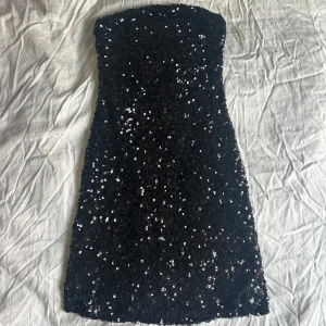 Svart paljettklänning - Snygg svart axelbandslös klänning täckt med glittriga paljetter. Klänningen har en figurnära passform och slutar vid mitten av låret. Den är endast använd en gång och är normal i storleken