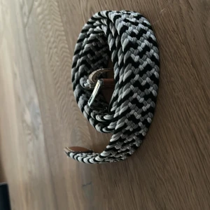Svartvitt flätat bälte - Snyggt flätat bälte i svart och vitt med ett stilrent mönster. Bältet har en silverfärgad spänne och är perfekt för att ge en extra touch till din outfit. Passar bra till både jeans och chinos.