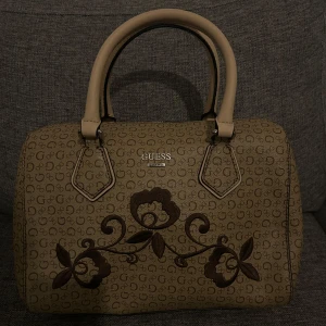 Beige handväska från Guess - Snygg beige handväska från Guess med brunt blommönster och logotyp. Väskan har två handtag och en dragkedja upptill. Perfekt för att ge en stilren touch till din outfit.