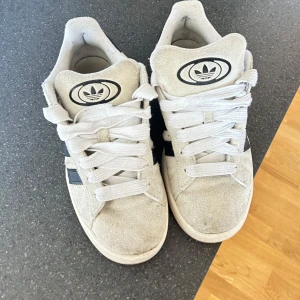 Adidas campus skor - Säljer ett par Adidas Campus sneakers i beige mocka med svarta detaljer och vita snören. Skorna har den klassiska tre-randiga designen på sidorna och en gummisula. Perfekta för en avslappnad stil.
