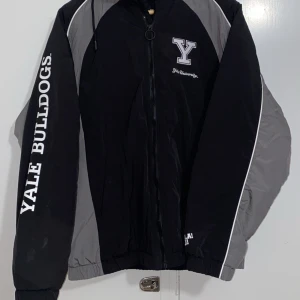 Svart och grå Yale Bulldogs jacka - Säljer en cool svart och grå jacka med Yale Bulldogs tryck. Jackan har dragkedja och långa ärmar, perfekt för höst och vår. Den är i bra skick och har en sportig stil med universitetstema. Perfekt för dig som vill ha en unik och bekväm jacka!